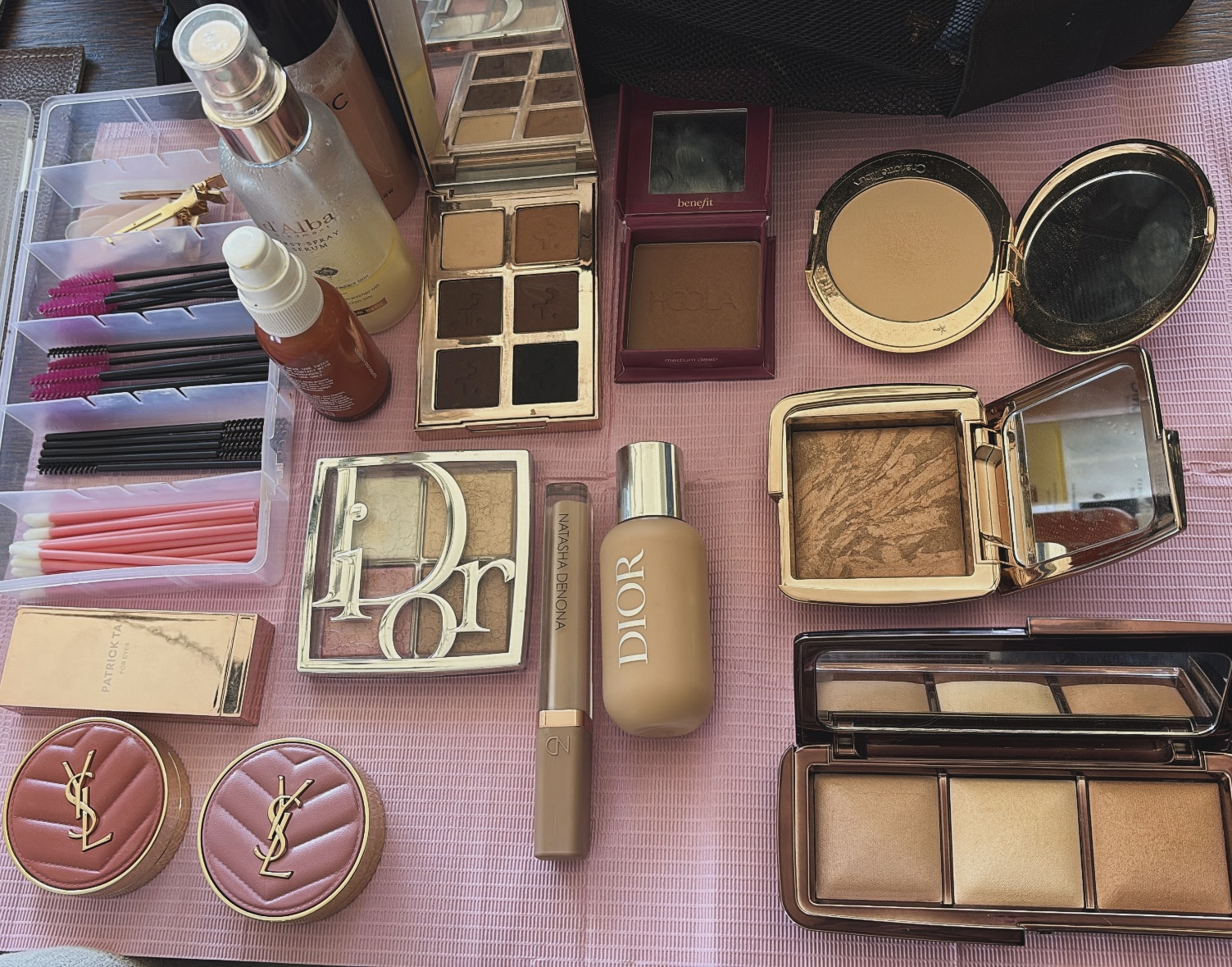 Makeup products including eyeshadow palettes, foundation, blush, lip gloss, and brushes arranged on a pink surface.