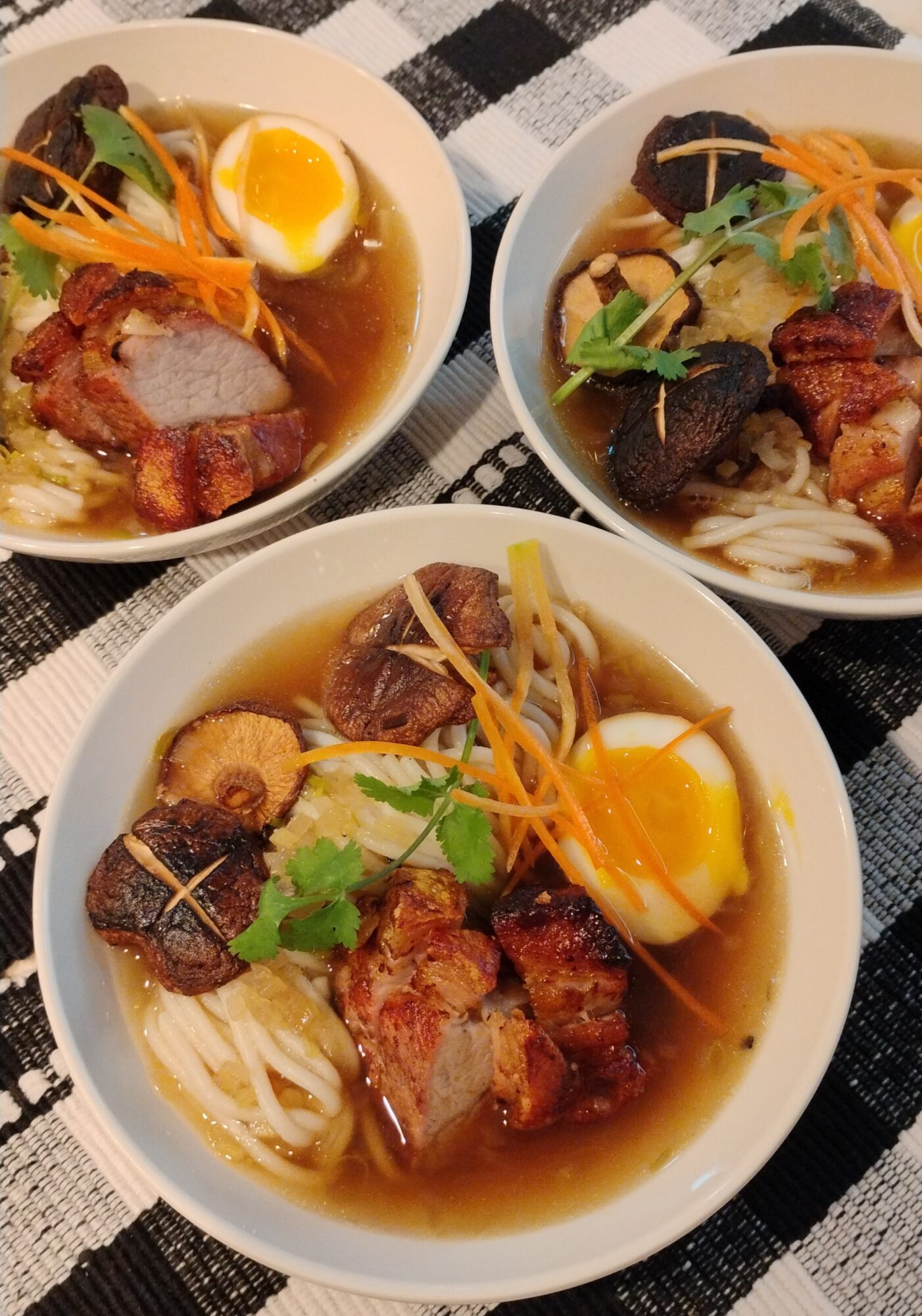 Three bowls of noodle soup with boiled eggs, meat, vegetables, and garnishes on a checkered cloth.