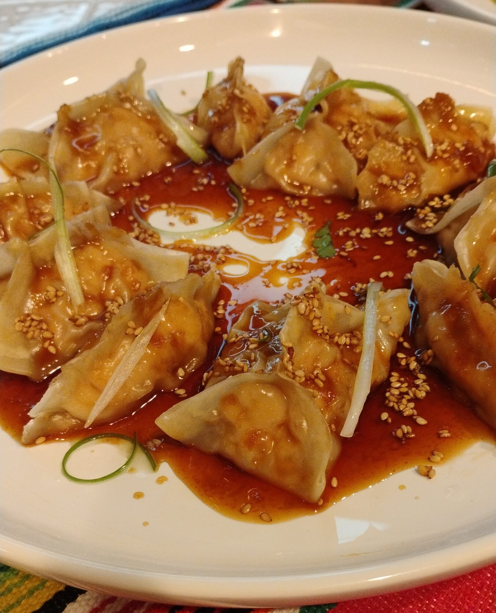 Plate of dumplings with sauce, garnished with sesame seeds and green onion slices, on a white dish.
