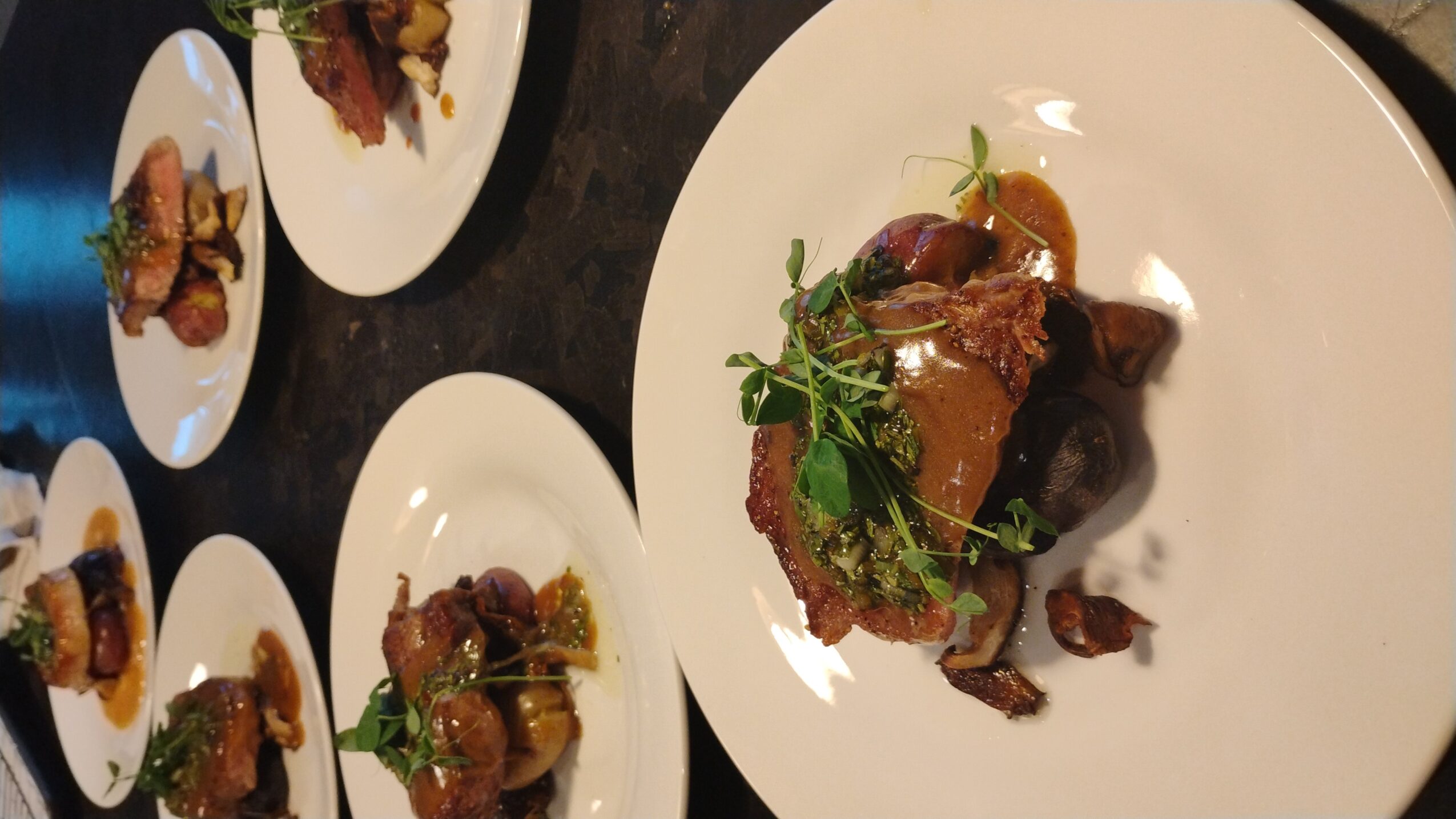 Multiple plates with gourmet food, including a main dish with meat, greens, and sauce, on a dark table surface.
