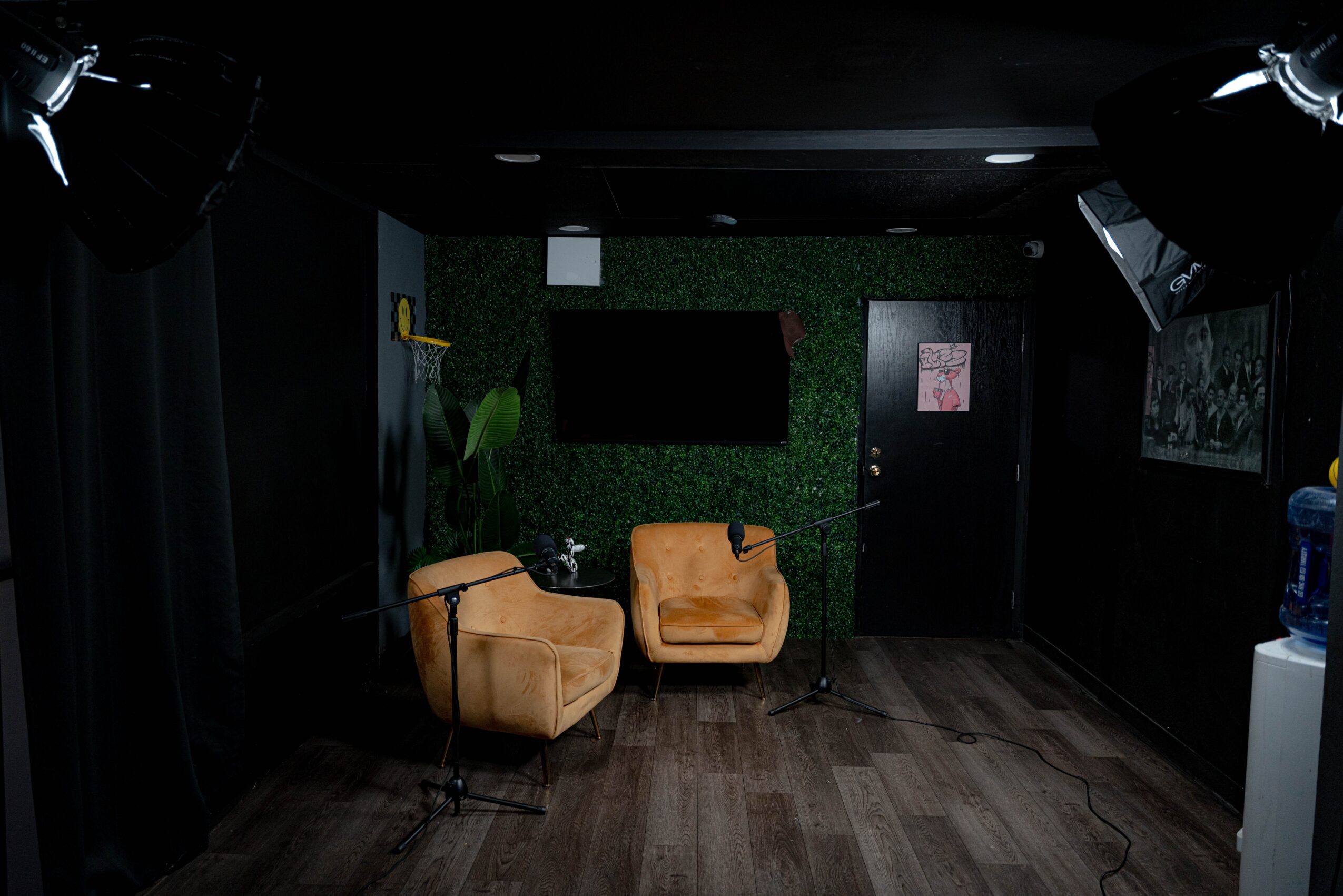 Room with two tan armchairs, dark walls, green wall with a black rectangle, and a door, wooden floor, and lighting equipment.
