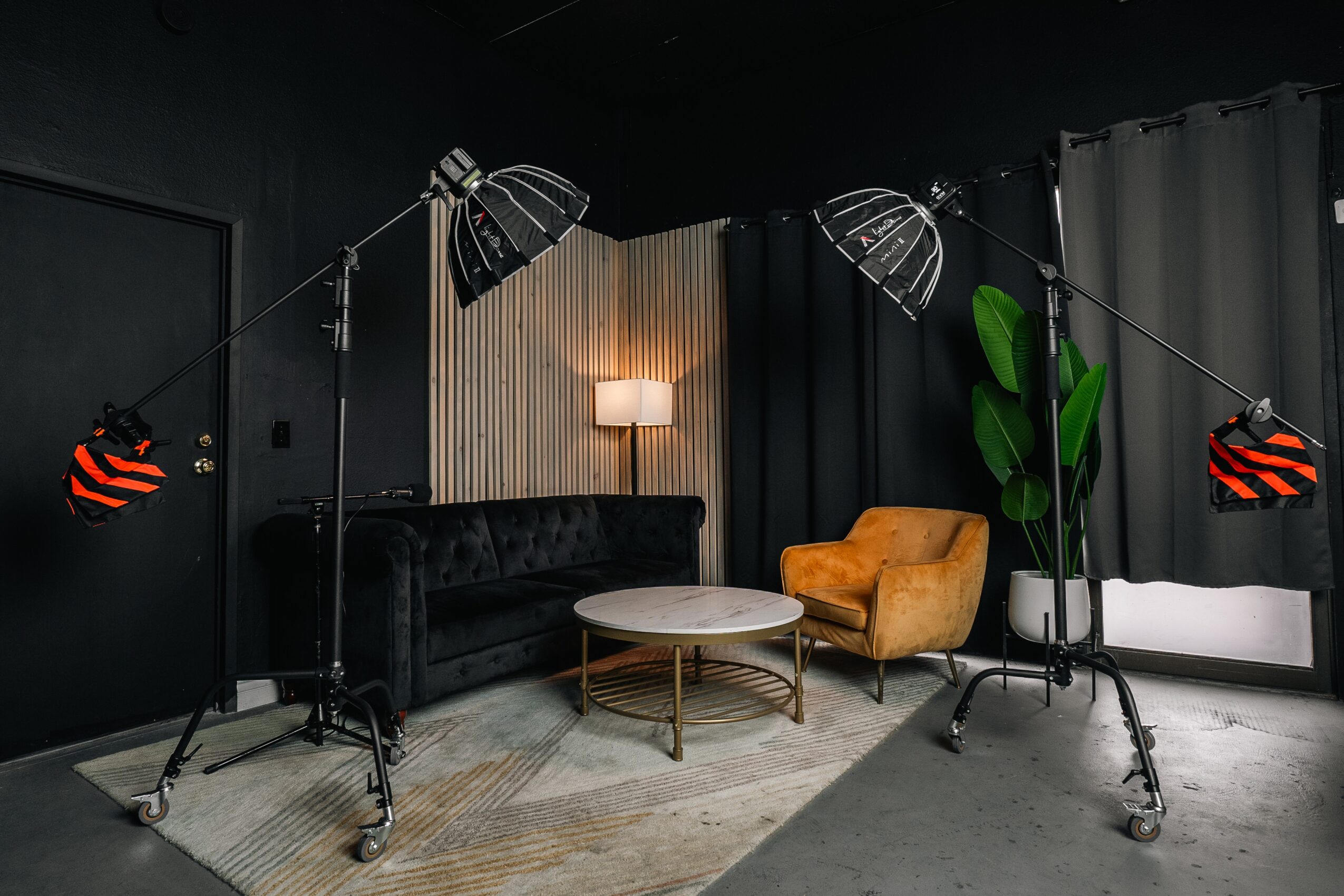 Studio with black sofa, yellow armchair, round coffee table, large plant, and lighting equipment, against dark walls and curtains.