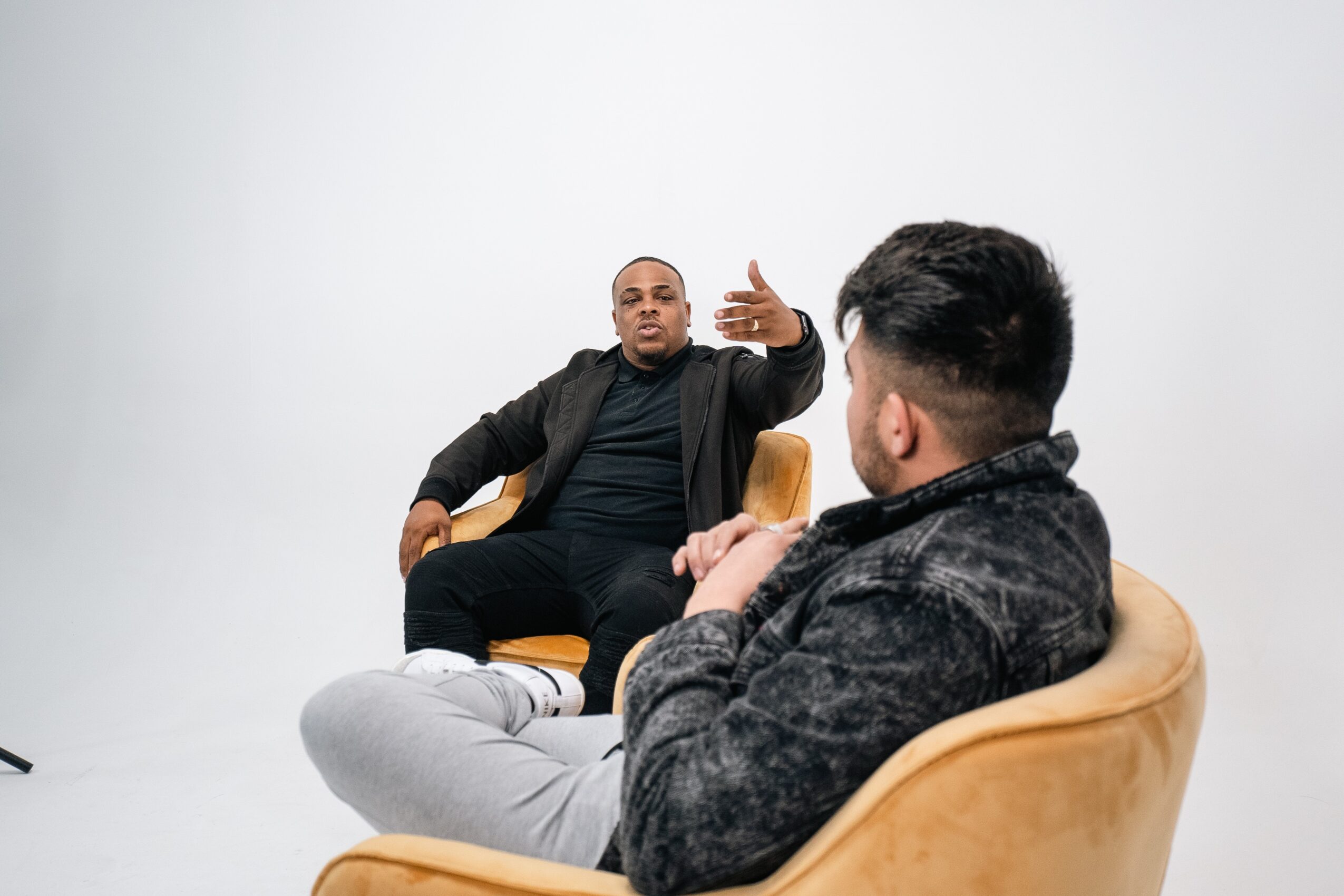 Two men engaged in conversation, one gesturing with hand, seated in chairs against a plain white background.