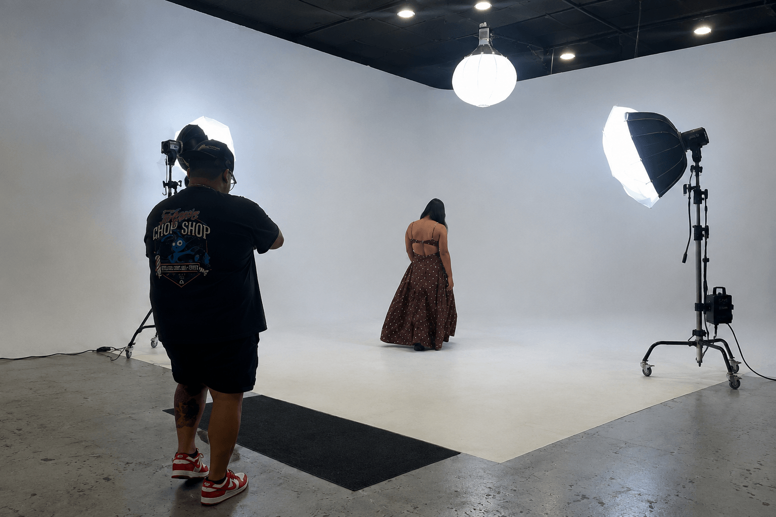 Person in black shirt and shorts filming a woman in a long dress in a photography studio with lighting equipment