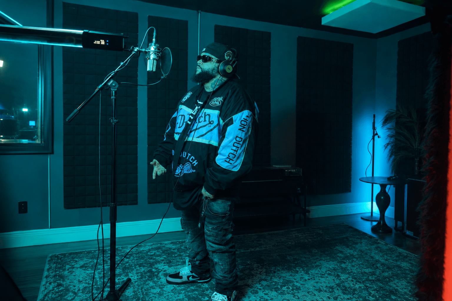 Person wearing headphones and sunglasses stands in a recording studio with a microphone, dark walls, and blue lighting.