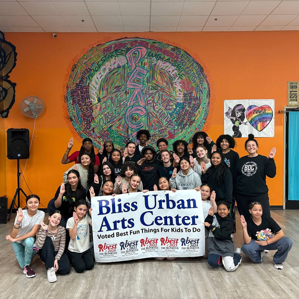 Group of children and adults holding a banner in front of a colorful mural, celebrating Bliss Urban Arts Center.