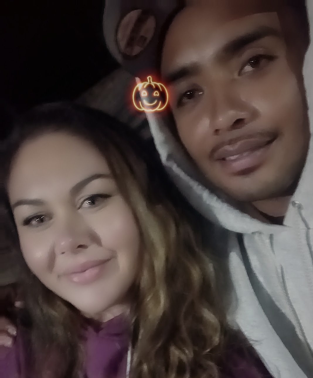 Two people lying close together, smiling, with a pumpkin-shaped light filter above them, in a dark setting.