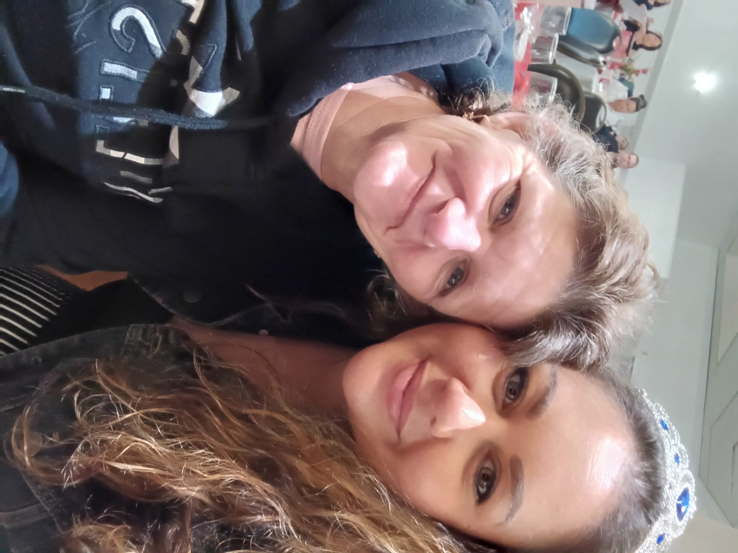 Two women smiling, one with curly hair and the other with straight hair, in a crowded indoor setting.