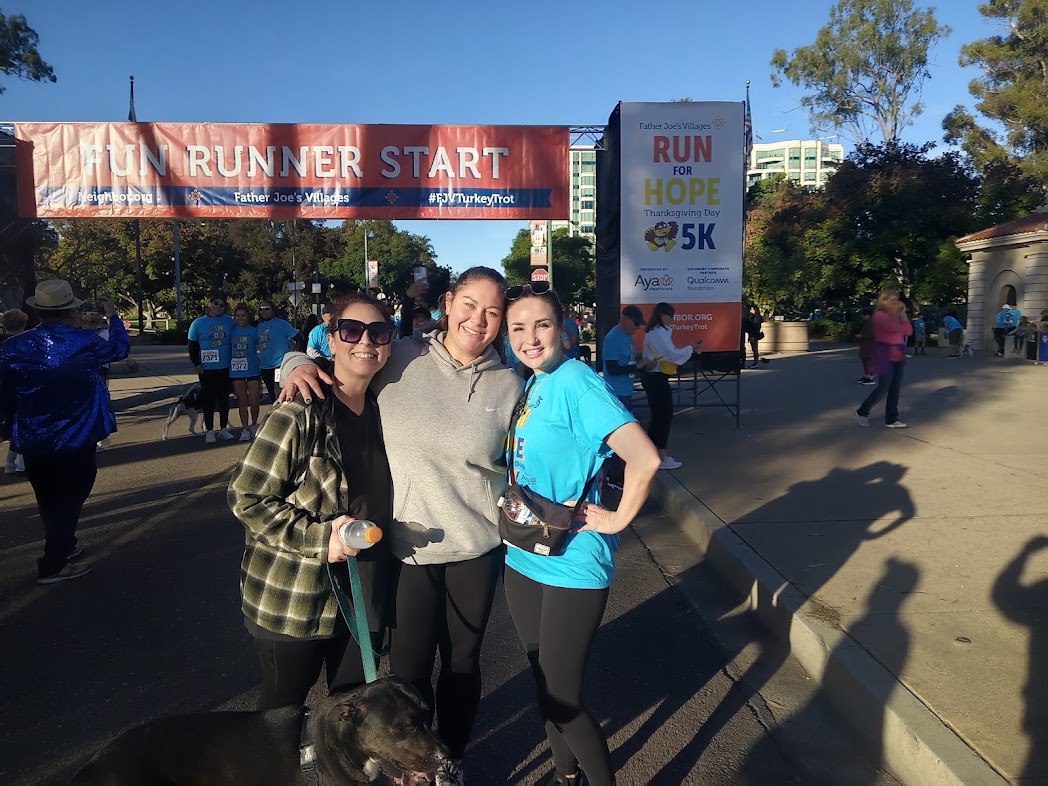 Three women smiling and posing together outdoors at a running event, with a banner and other participants in the background.