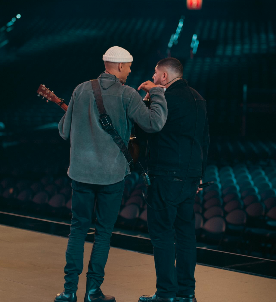 Two men standing on a stage, one holding a guitar, engaged in conversation, with empty seats in the background.