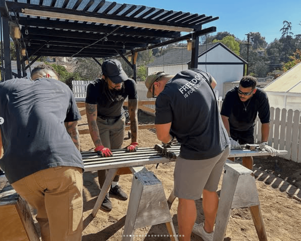 Four people work together on a metal project outdoors under a shelter, with a white fence and trees in the background.