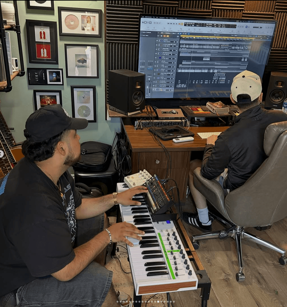 Two people in a music studio working on a digital audio workstation, one playing a keyboard, the other at a computer screen.
