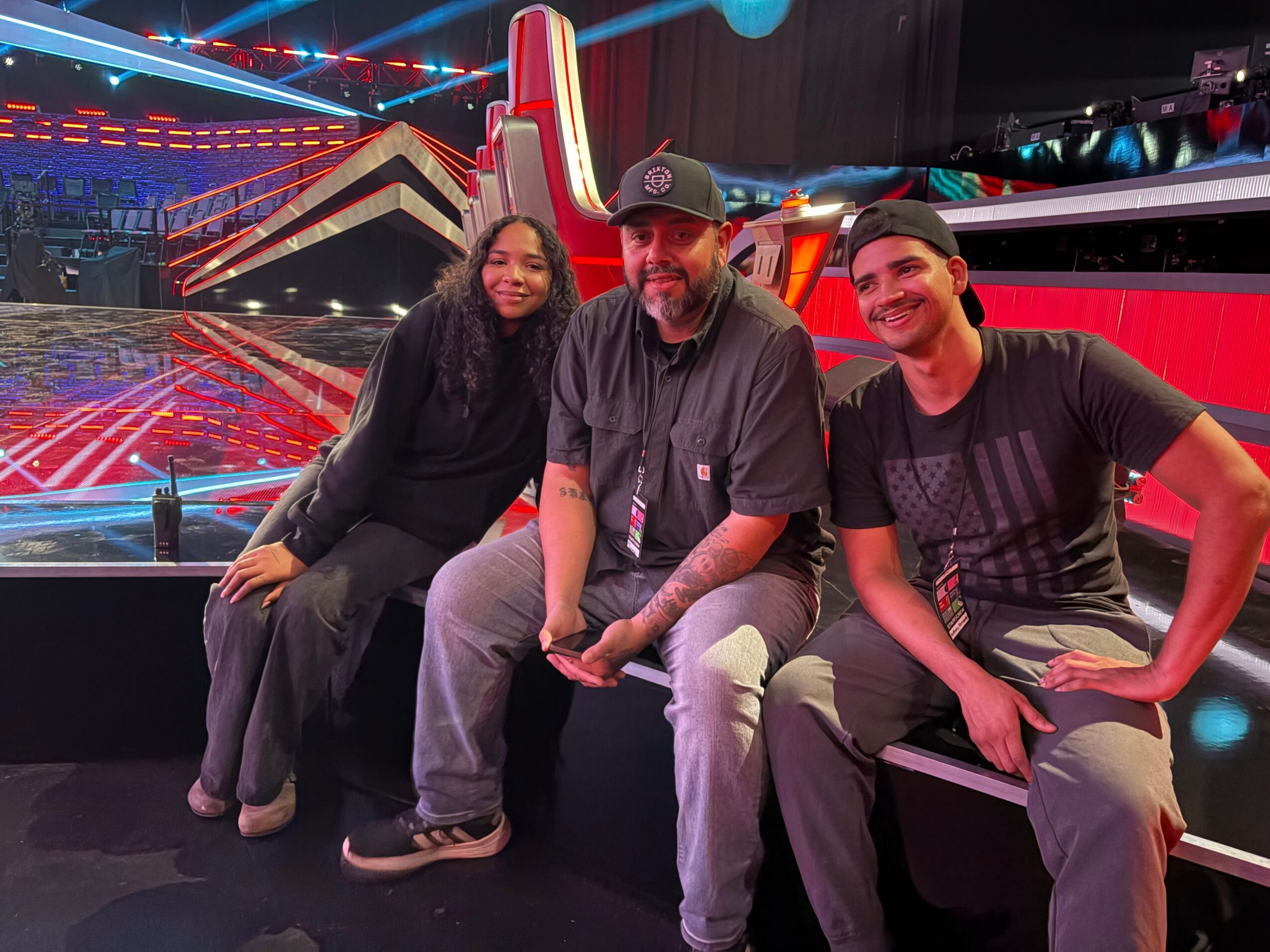 Three people sitting together in front of a colorful stage with bright lights and a large screen, smiling at the camera.
