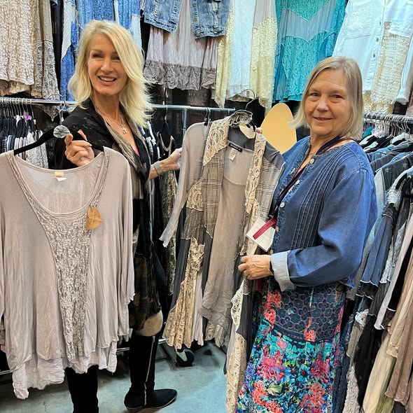 Two women shopping for clothes in a store, surrounded by hanging garments, smiling and holding clothing items.