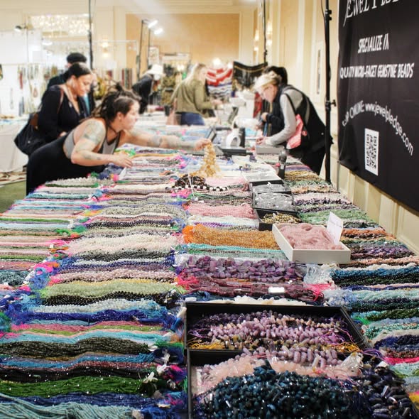 People browsing colorful jewelry and accessories displayed on tables at an indoor market or event.