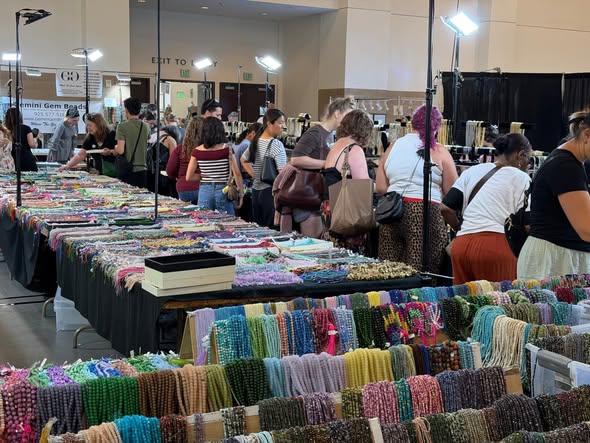 People browse colorful handmade jewelry and accessories at a market stall indoors.