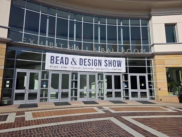 Entrance of a building with a banner for the Bead & Design Show, glass doors, and brick pavement.