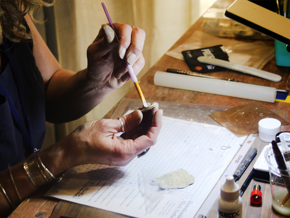 Person's hands working with a small object, surrounded by art supplies on a wooden table, with papers and tools visible.