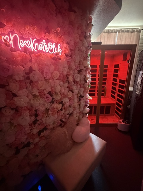 A person with a fluffy, textured garment stands in front of a red-lit sauna or infrared cabin, with a window showing the interior.