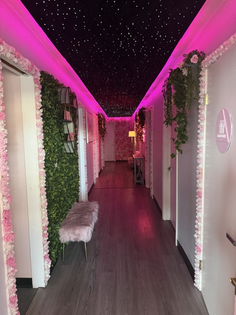 Decorated hallway with pink lighting, floral arrangements, and a fluffy stool, leading to a door at the end.