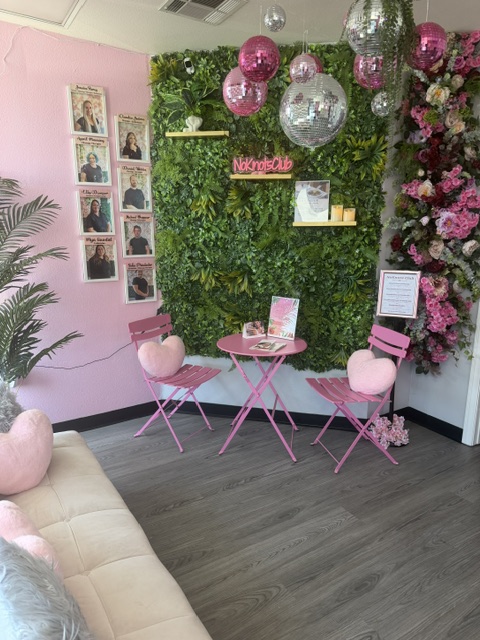 Decorative pink and white chairs with balloons, pink flowers, and framed photos on a pink wall, with a green plant backdrop.