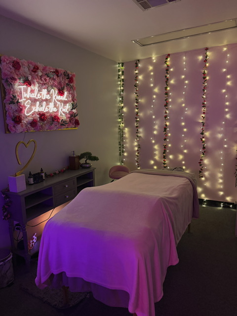 Room with a bed, fairy lights on wall, floral sign, and a small table with decorative items, dimly lit with purple and yellow lighting.