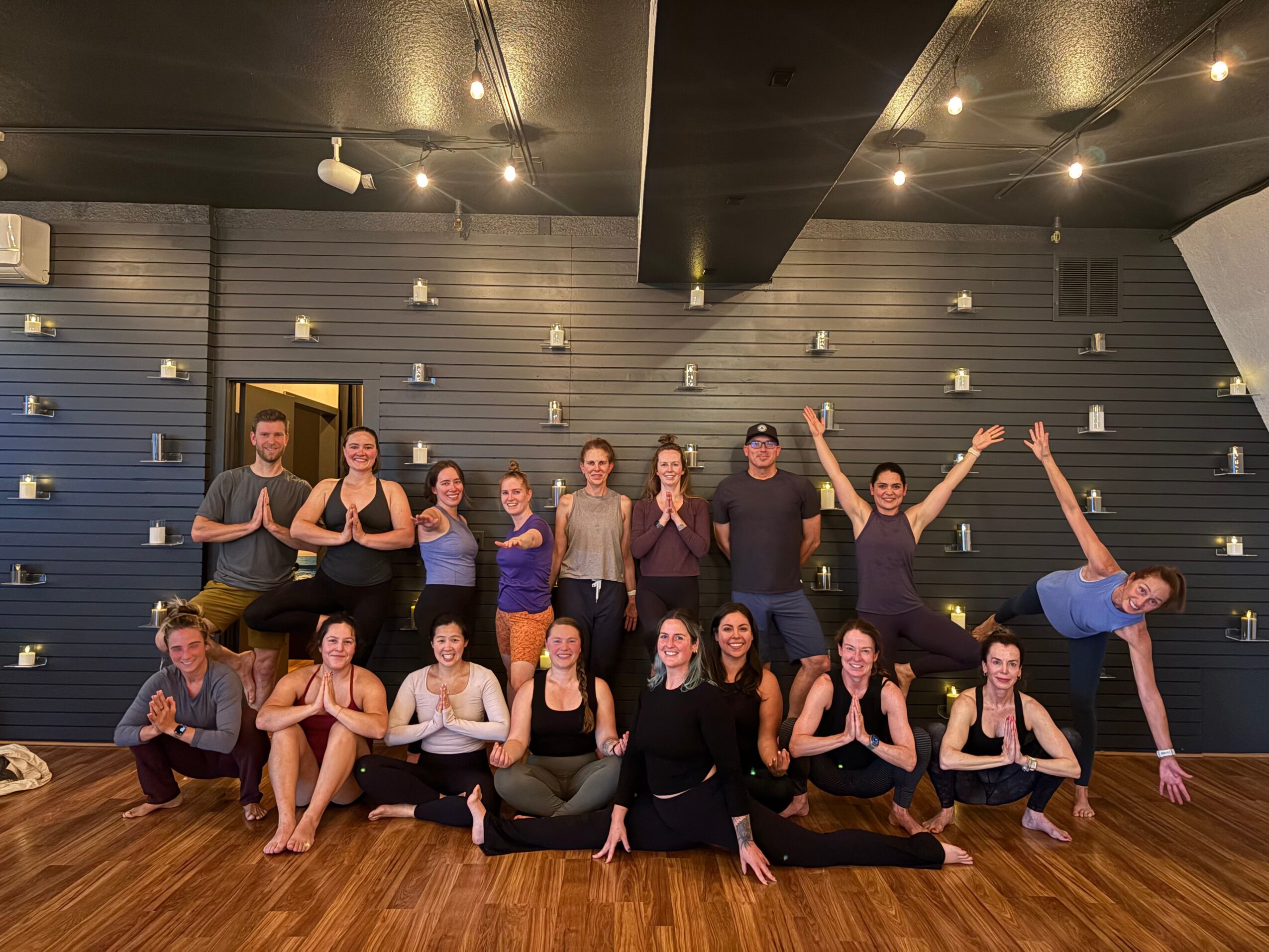 Group of 14 people in a dance studio, some sitting and some standing, posing with hands in prayer or raised, wooden floor, black wall, ceiling lights.