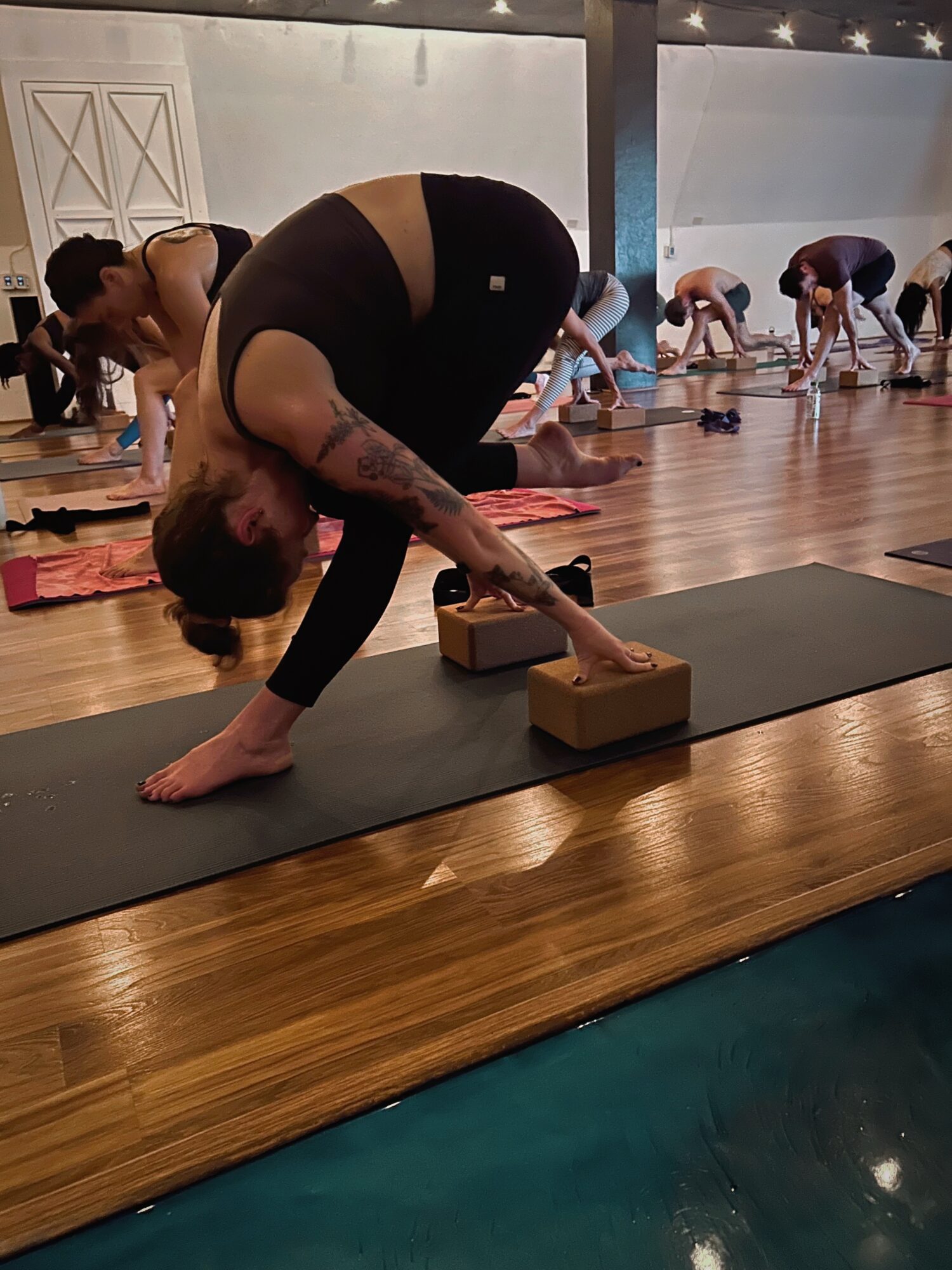 People practicing yoga in a studio, with a woman in foreground using yoga blocks, others in downward dog pose in background.