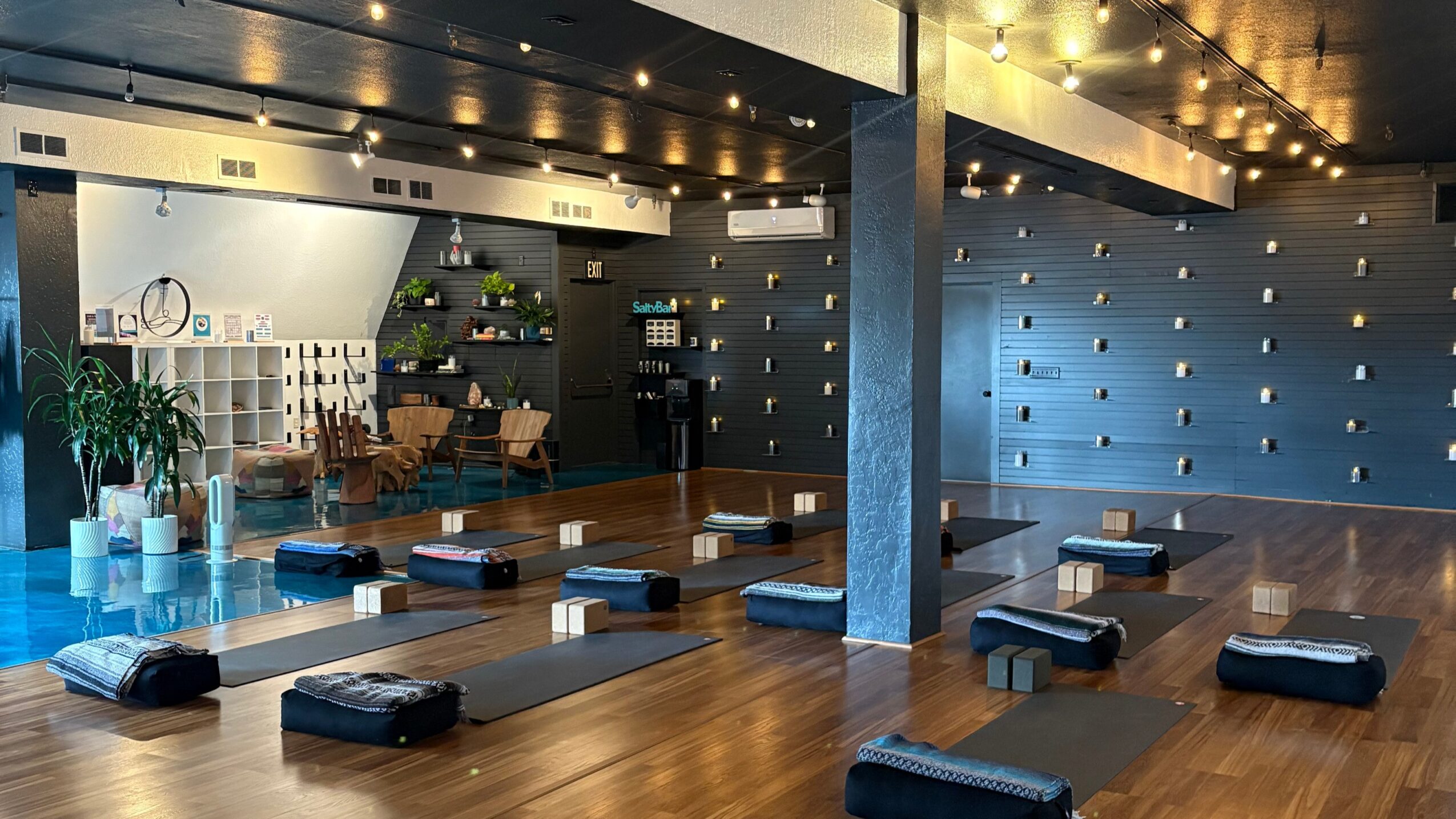 Yoga or meditation studio with cushions on wooden floor, lockers, and plants, illuminated by warm ceiling lights.