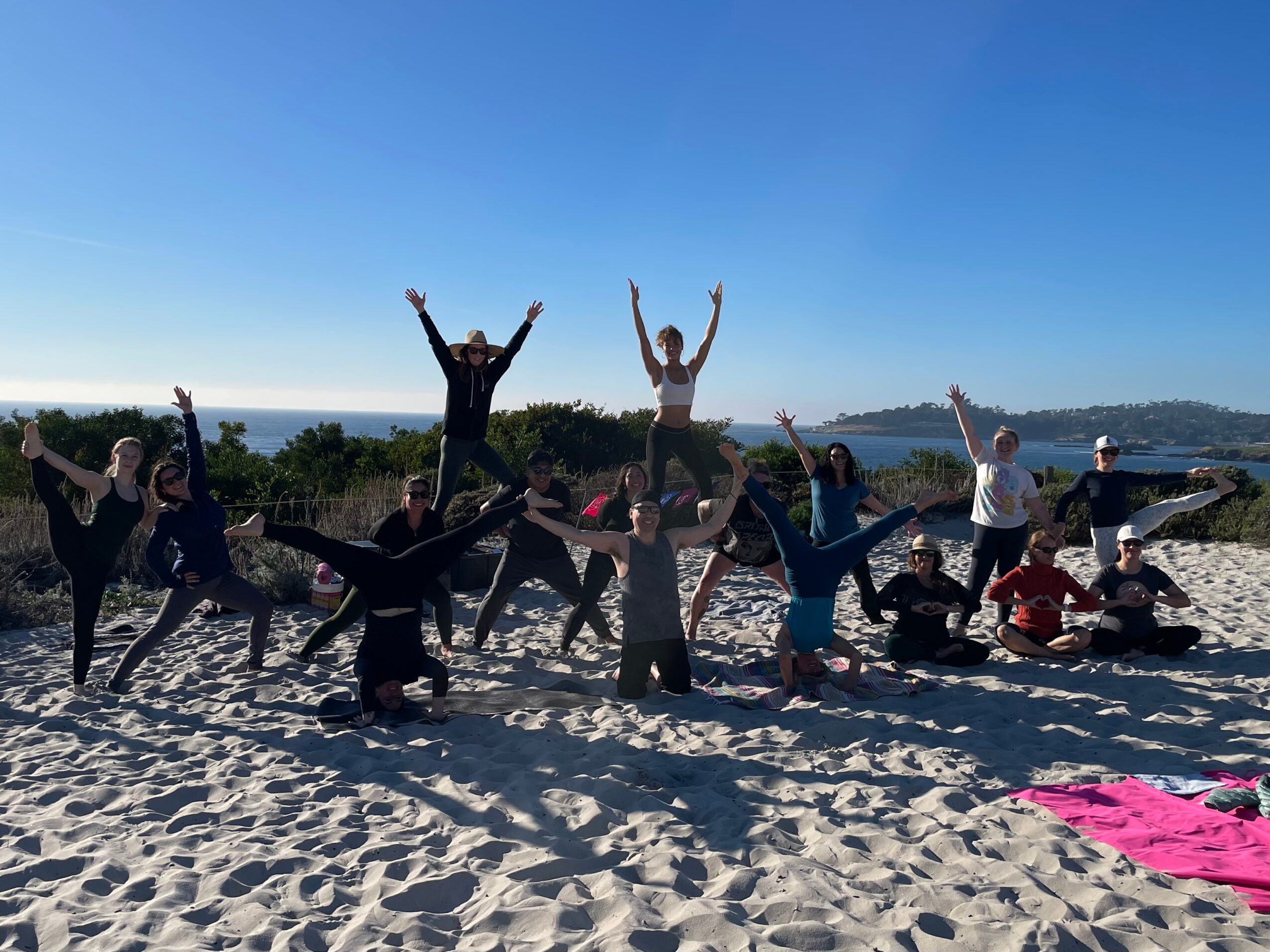 Group of people on a beach, some standing and some sitting, with arms raised, under a clear blue sky.