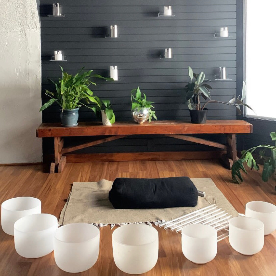 Indoor space with a wooden bench, plants, candles on a dark wall, and singing bowls on the floor, with a window on the right.