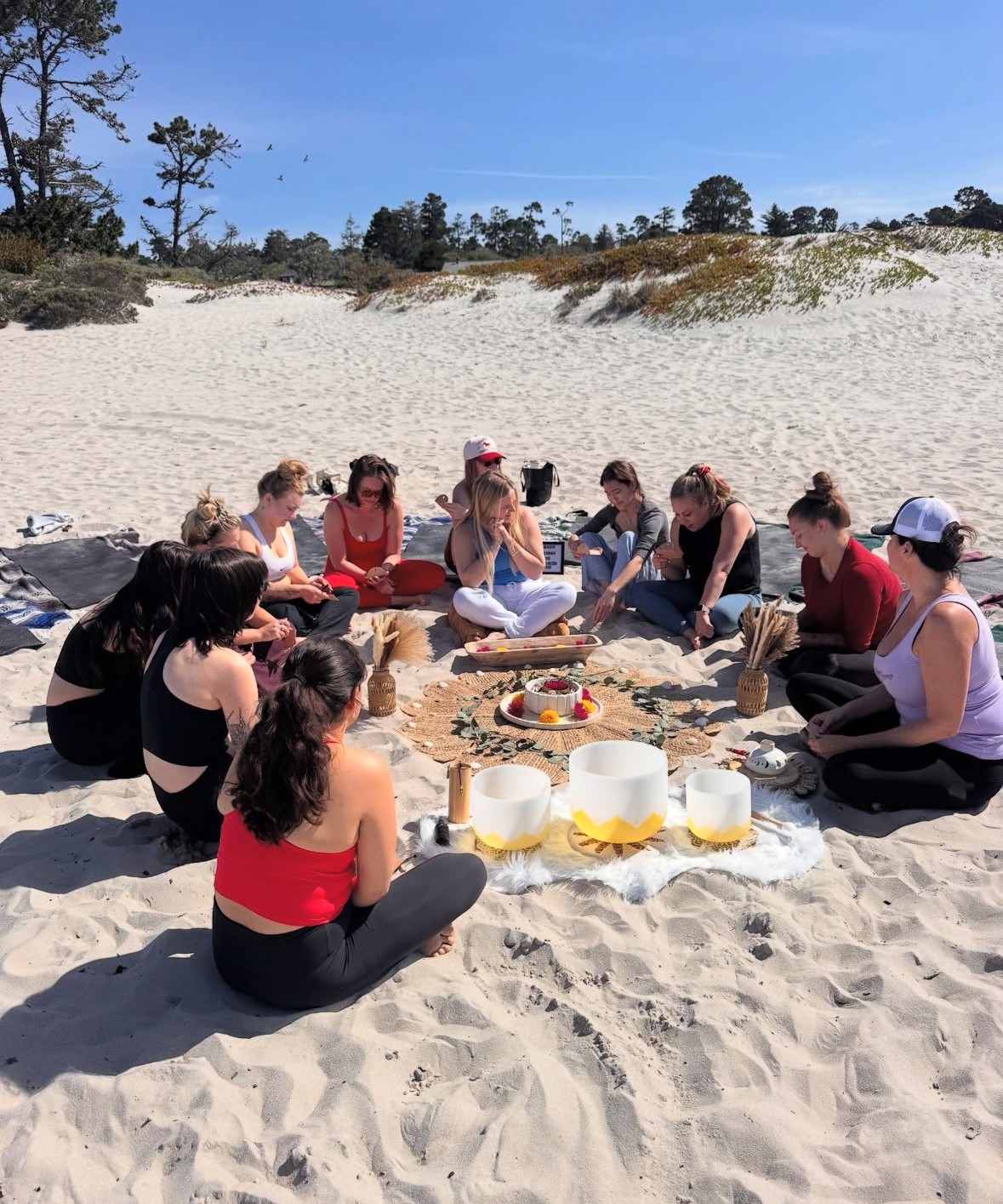 Group of people sitting in a circle on sandy beach, with food and drinks in the center, under a clear blue sky.