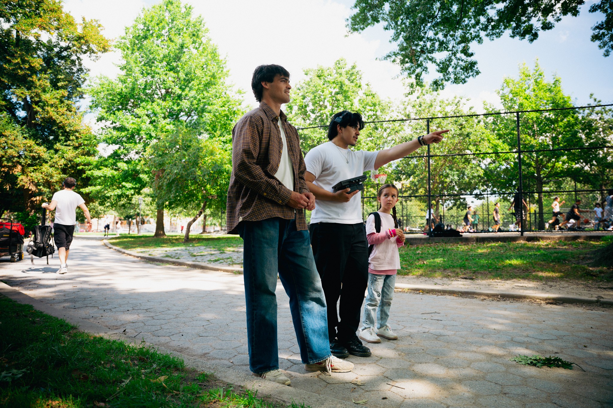 Three people standing on a park pathway, with trees and a fence in the background, one pointing at something.