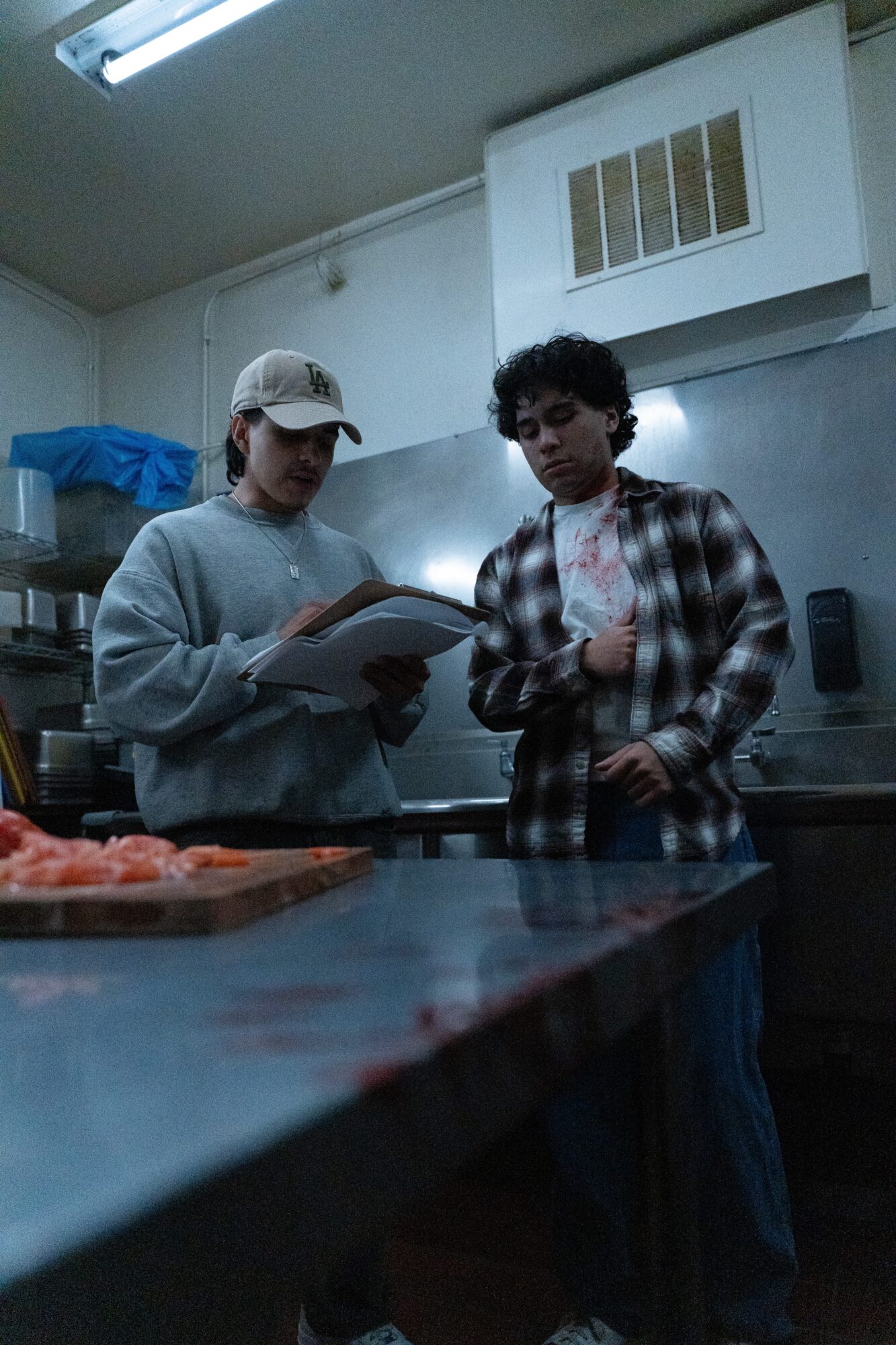 Two young men stand in a kitchen, one holding a notebook and the other looking at it, with food on the counter.