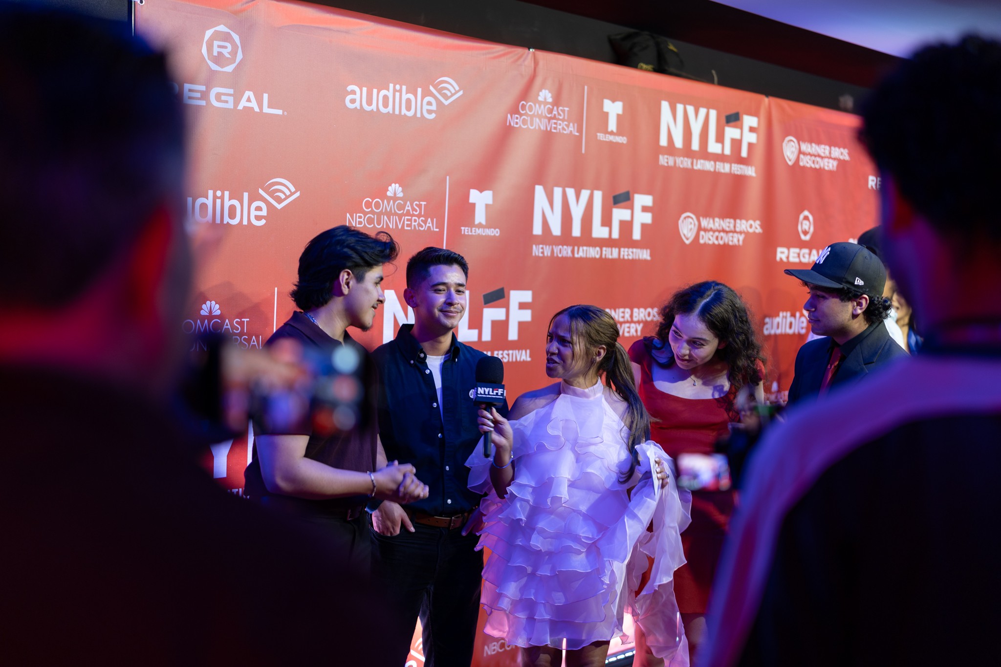 Group of people on red carpet with NYLFF backdrop, some holding microphones, engaging in conversation, dressed formally.