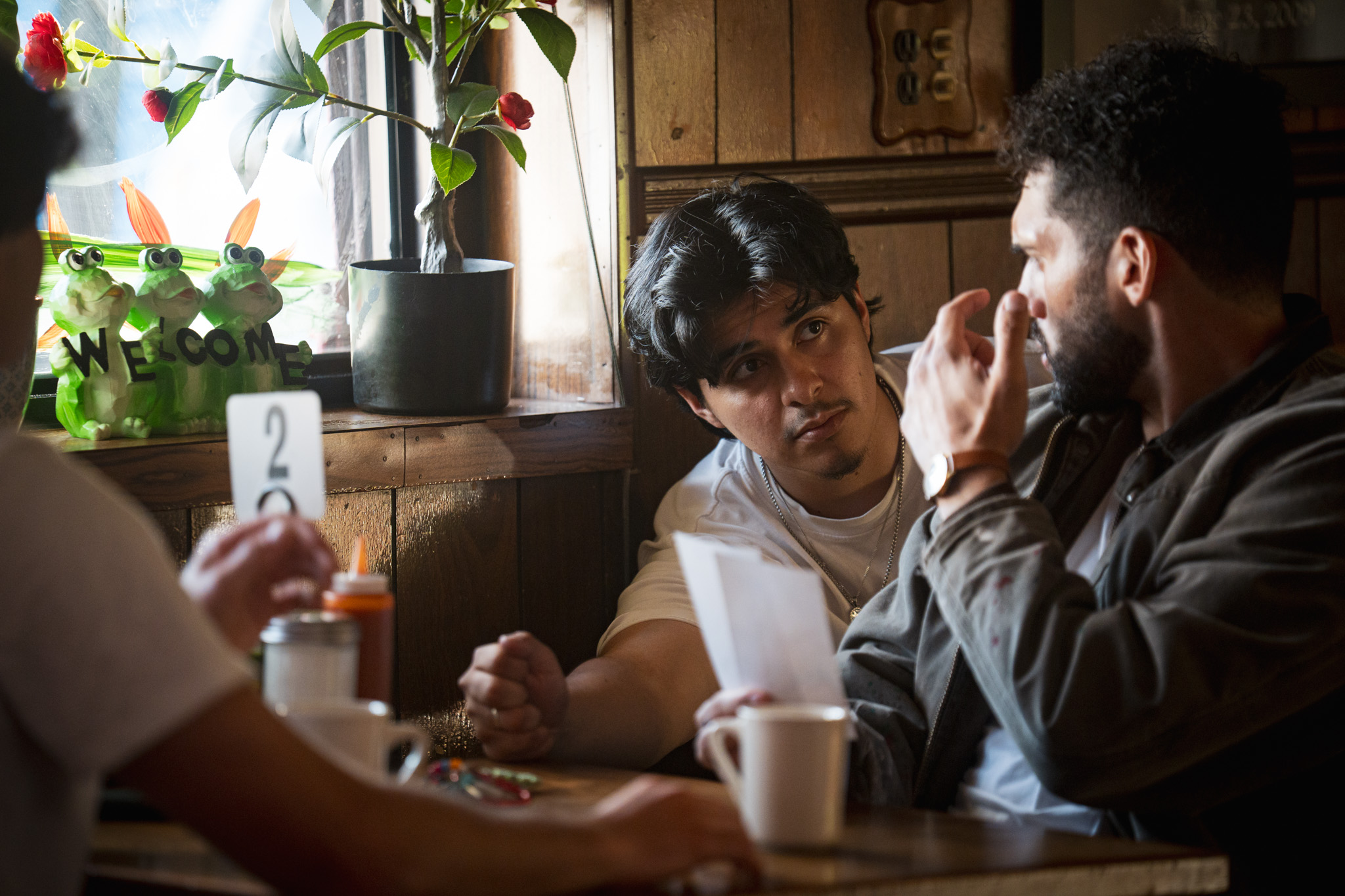 Two men sit at a table in a cozy cafe, engaged in conversation, with a window, plants, and a wooden wall in the background.