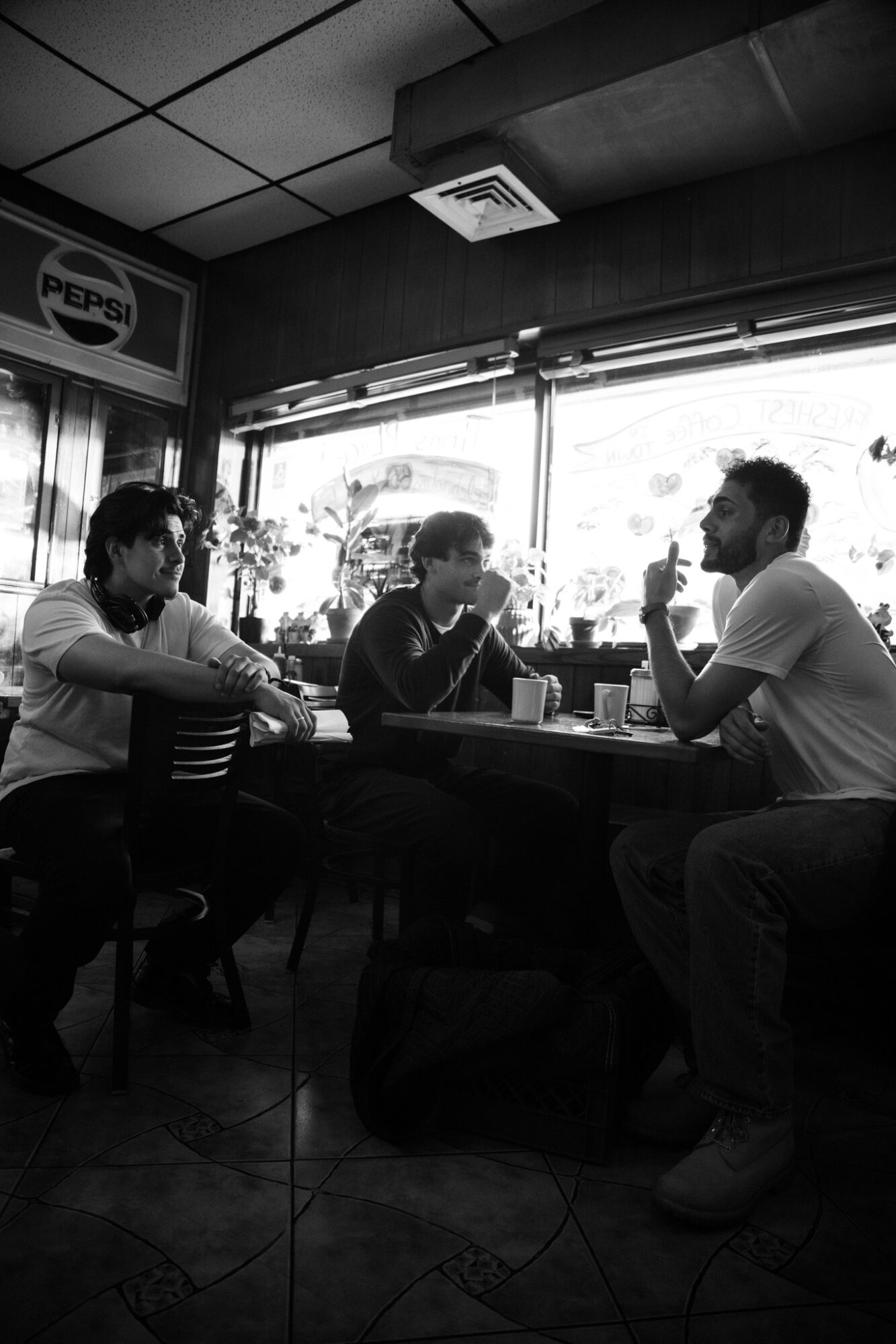 Three people sit at a table inside a cafe, engaged in conversation, with a window and Pepsi sign in the background.