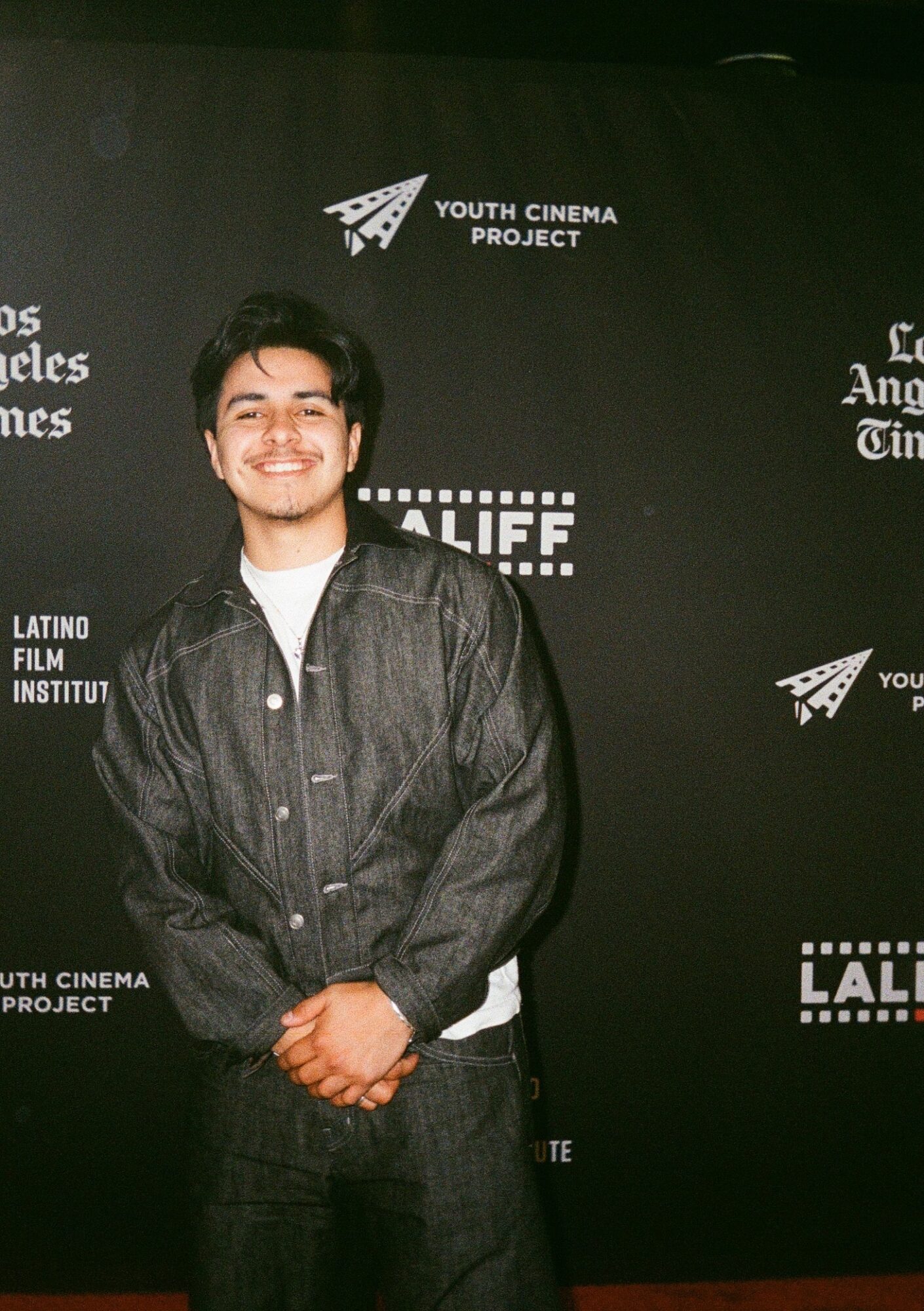 Young man smiling, standing in front of a black backdrop with logos and text, wearing a dark jacket and white shirt.