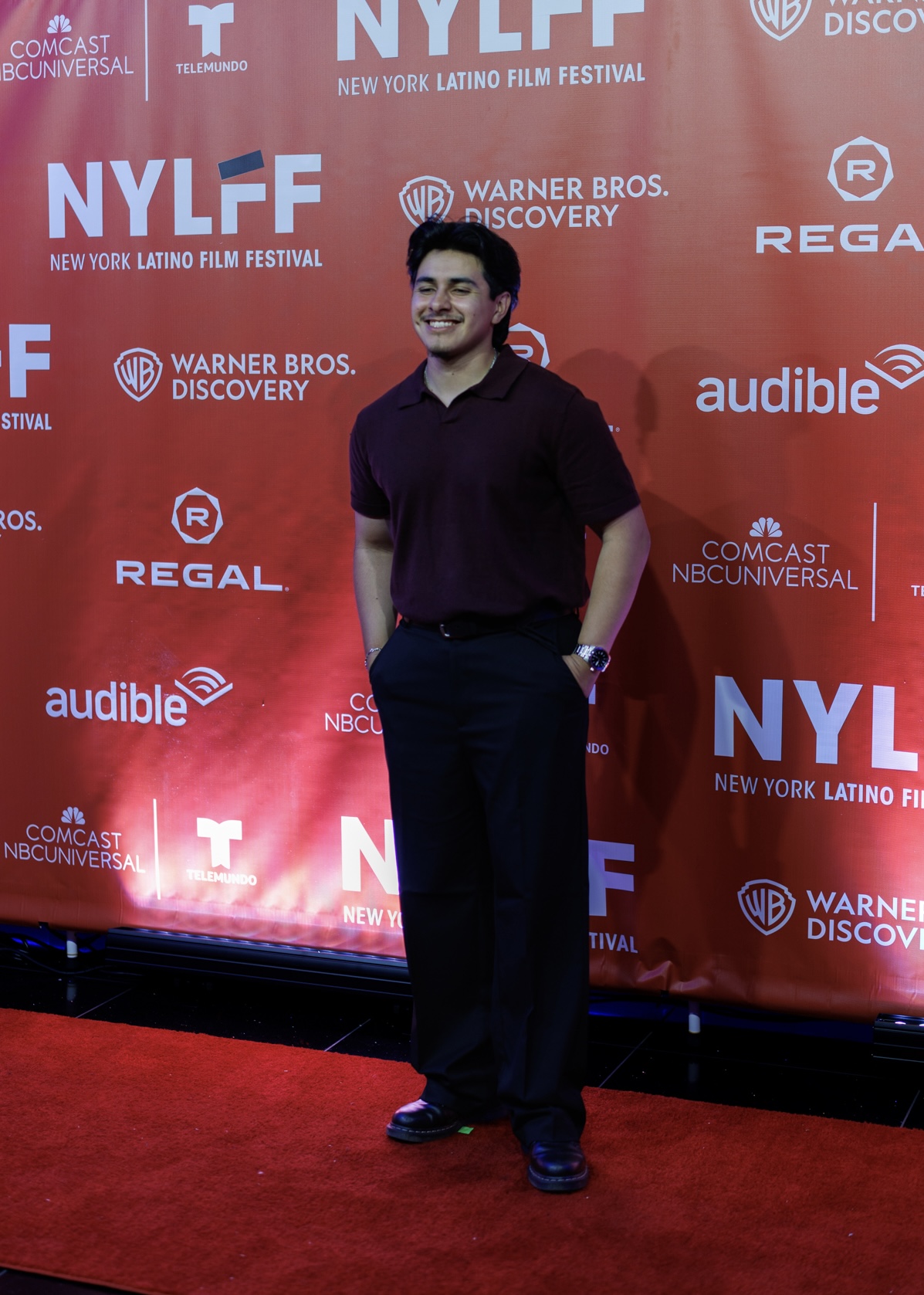 Man standing on red carpet in front of a red backdrop with event logos, smiling, hands in pockets, wearing a dark shirt and pants.