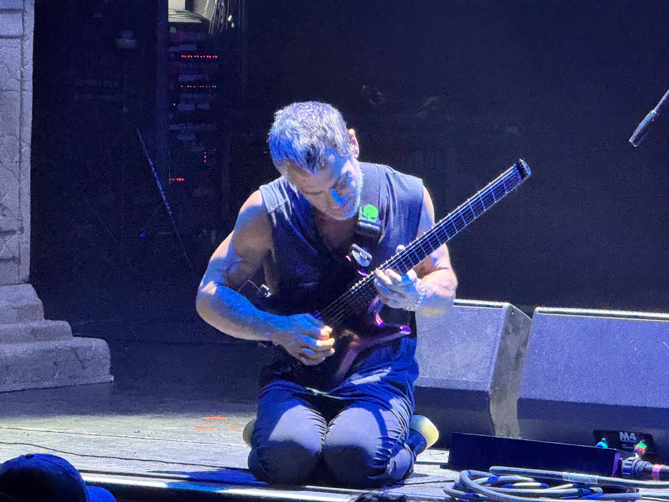 Person kneeling on stage playing a guitar, with stage equipment and dark background behind them.