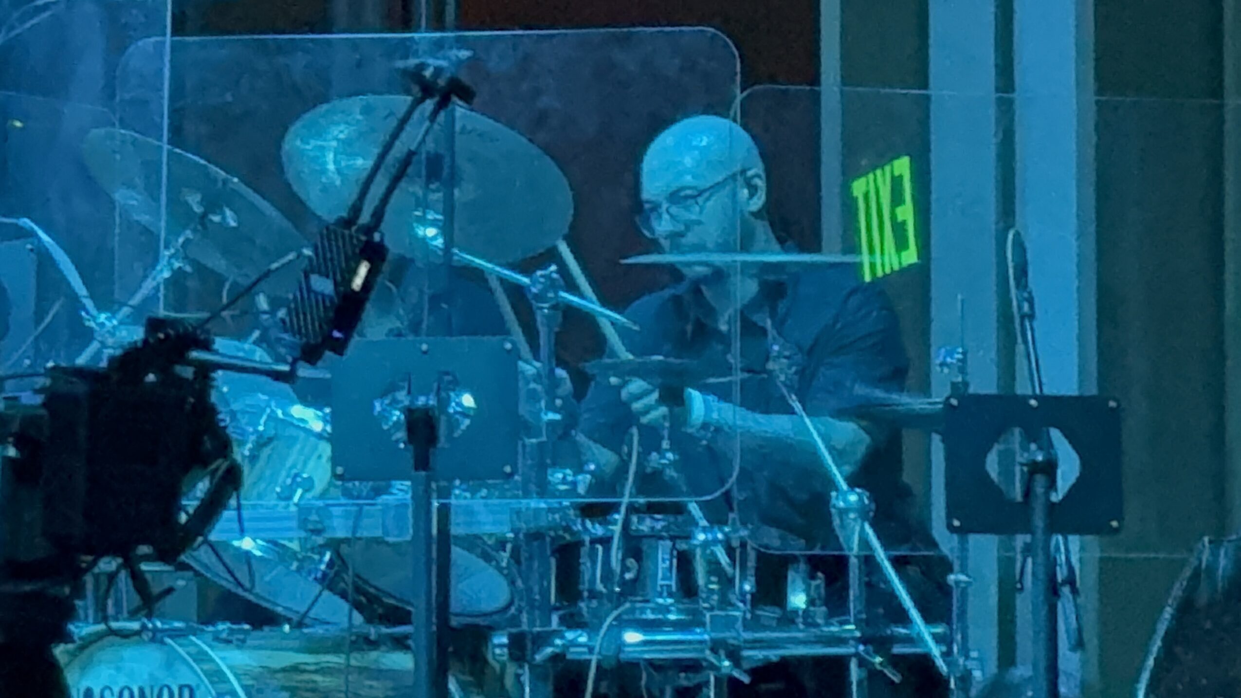 A person playing drums behind a transparent shield, with a green exit sign in the background.