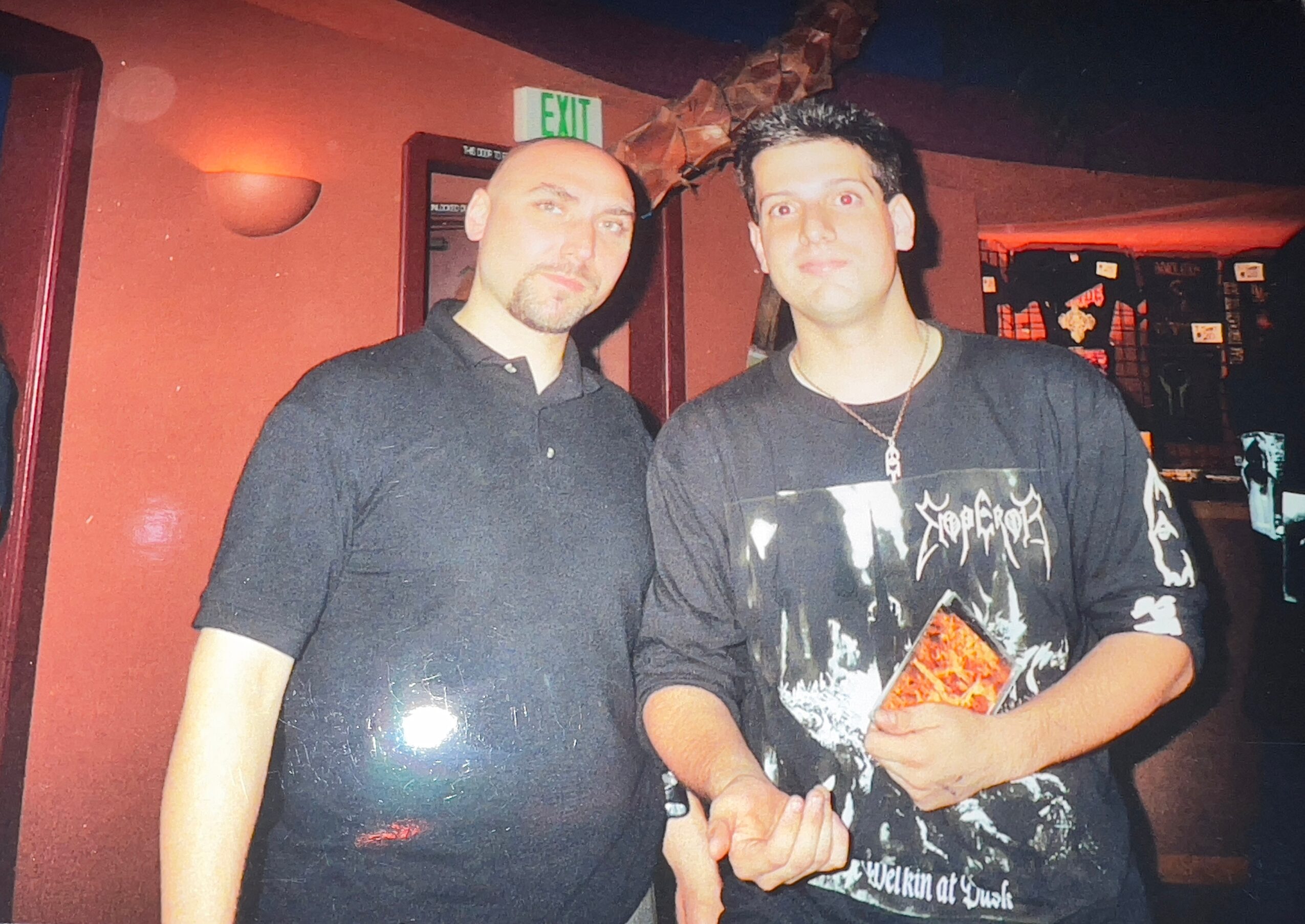Two men standing indoors, one holding a book or magazine, with a red wall and exit sign in background.