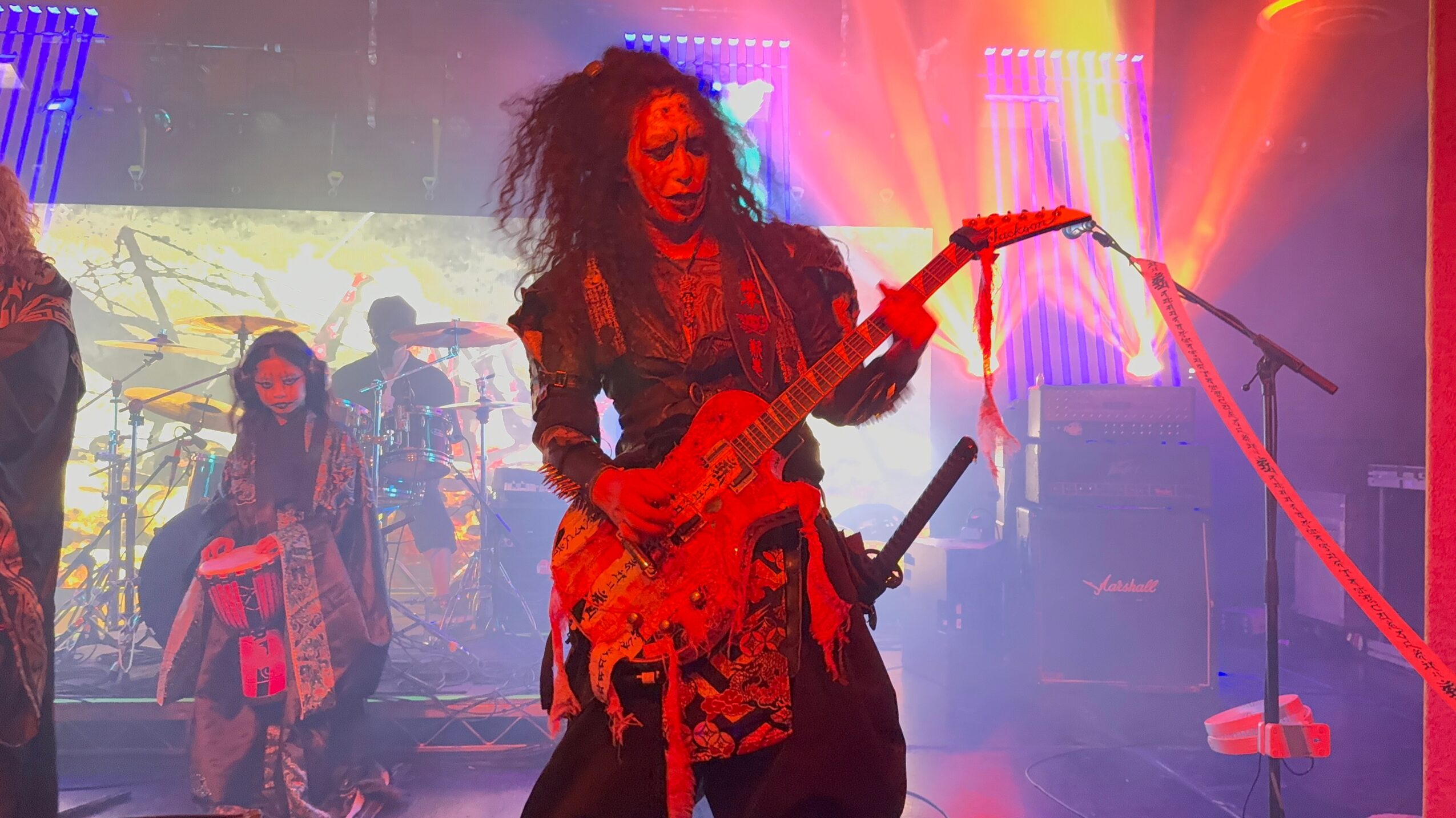 Female guitarist performing on stage with colorful lighting, other band members in background, vibrant atmosphere.