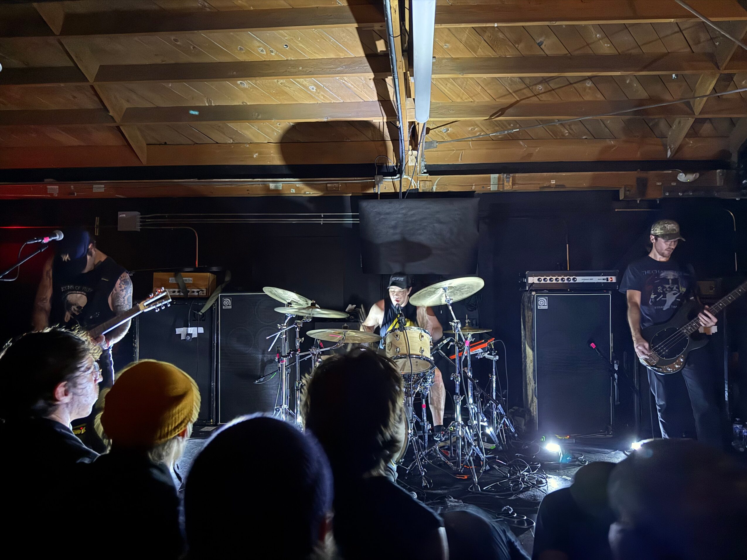 Band performs on stage with drums, guitars, and amplifiers, audience watching in a dimly lit venue with wooden ceiling.