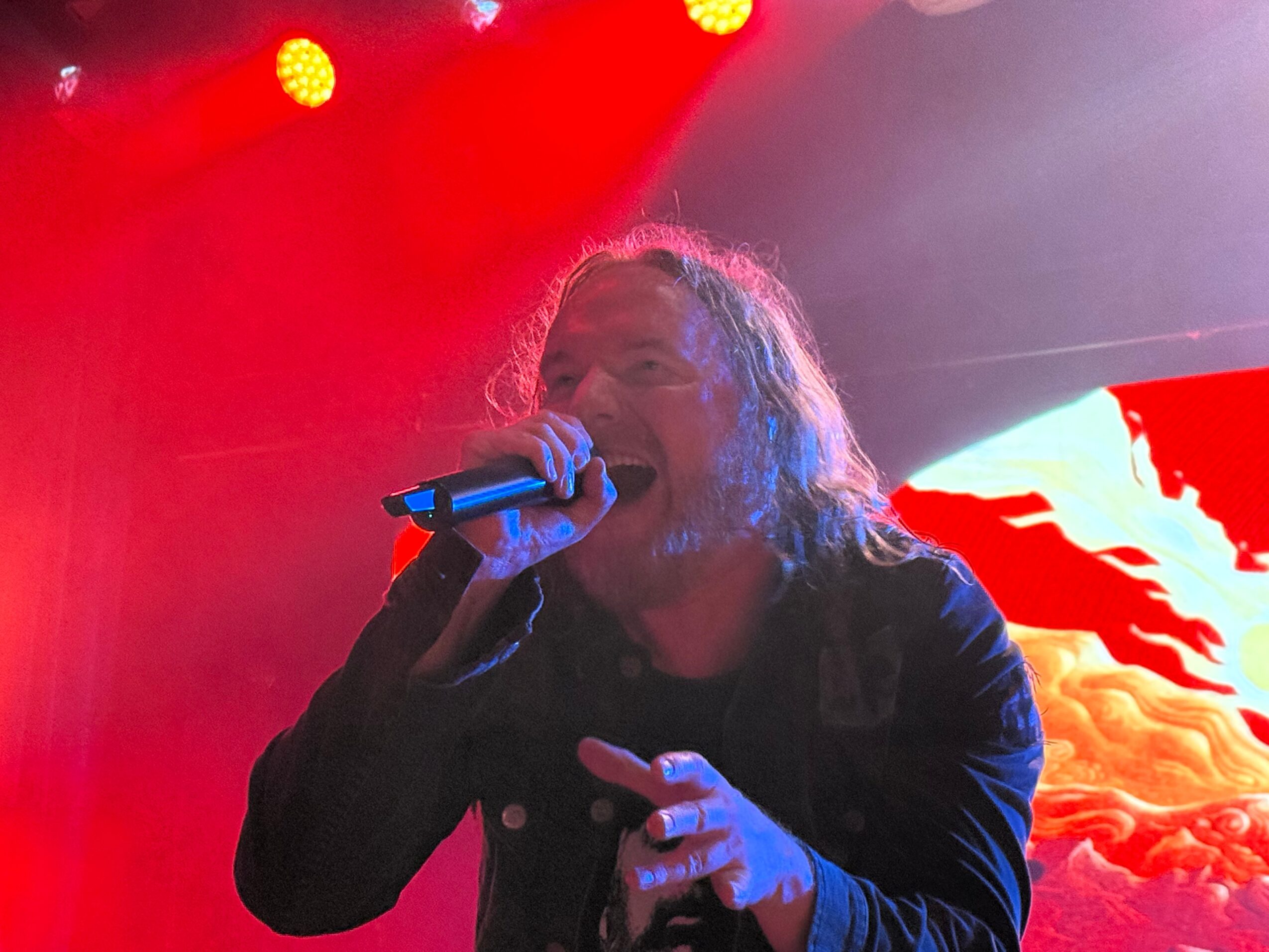 Person singing into microphone on stage with red and purple lighting, long hair, and a dark jacket.