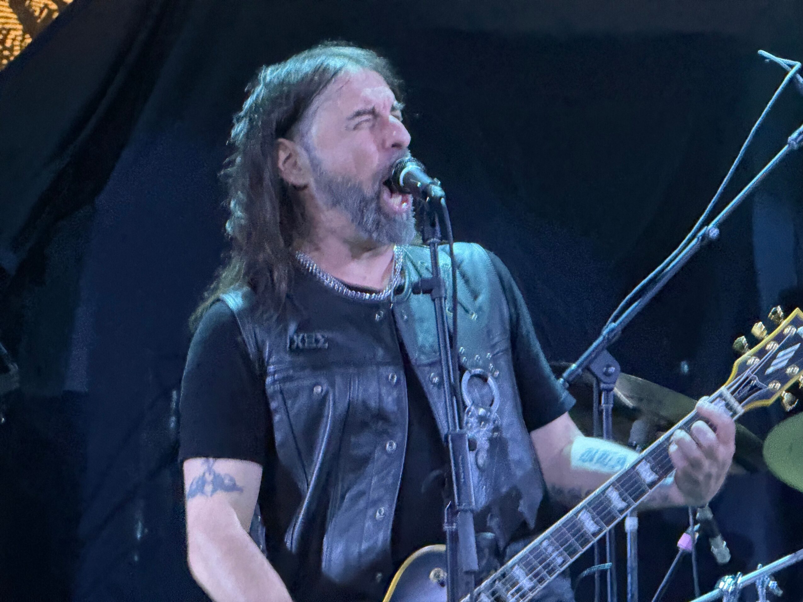 Man singing into microphone while playing guitar on stage, wearing a black vest and t-shirt, with long hair and beard.