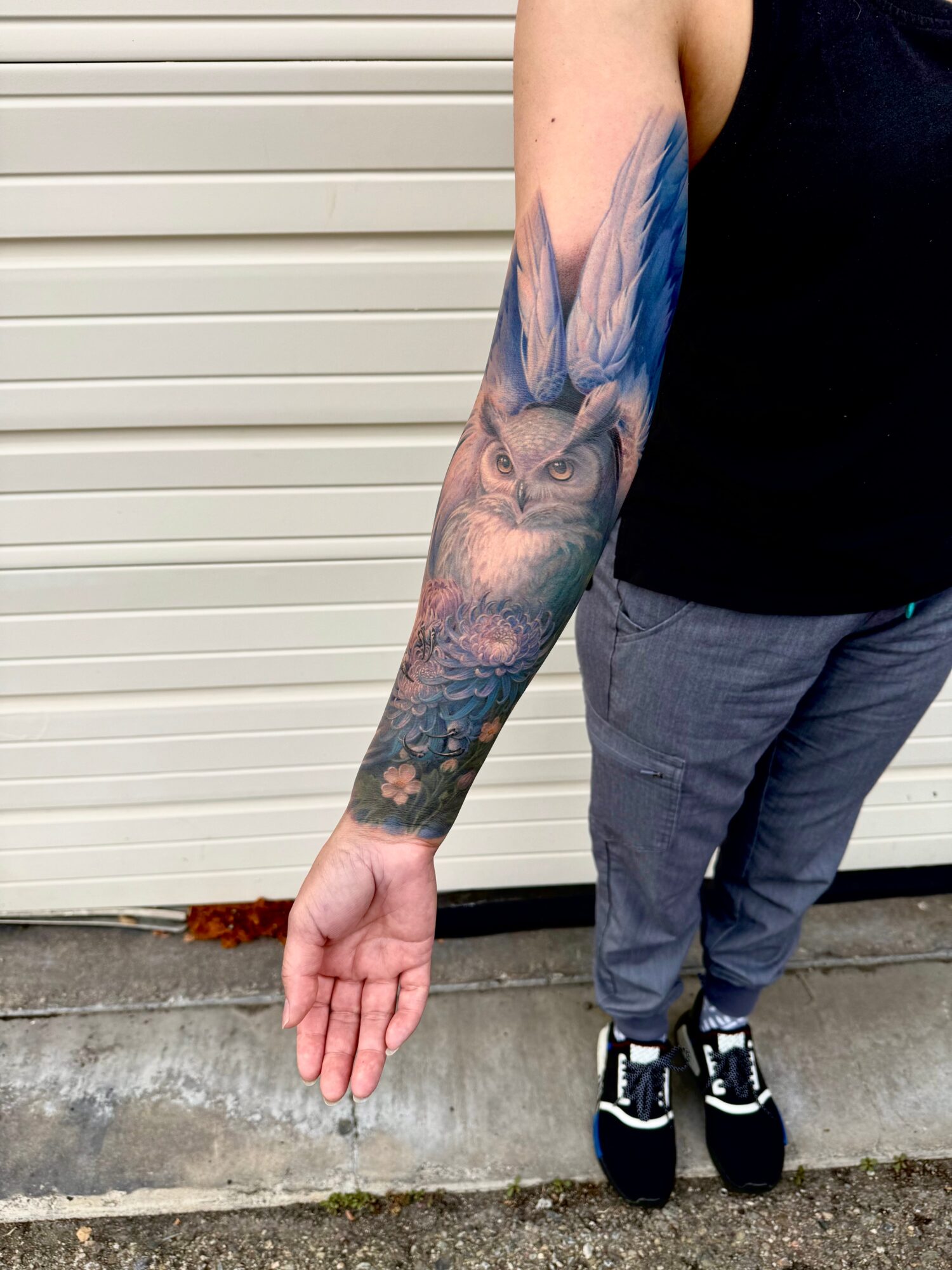 Person with a detailed owl tattoo on their arm, wearing a black sleeveless top and gray pants, standing outdoors.