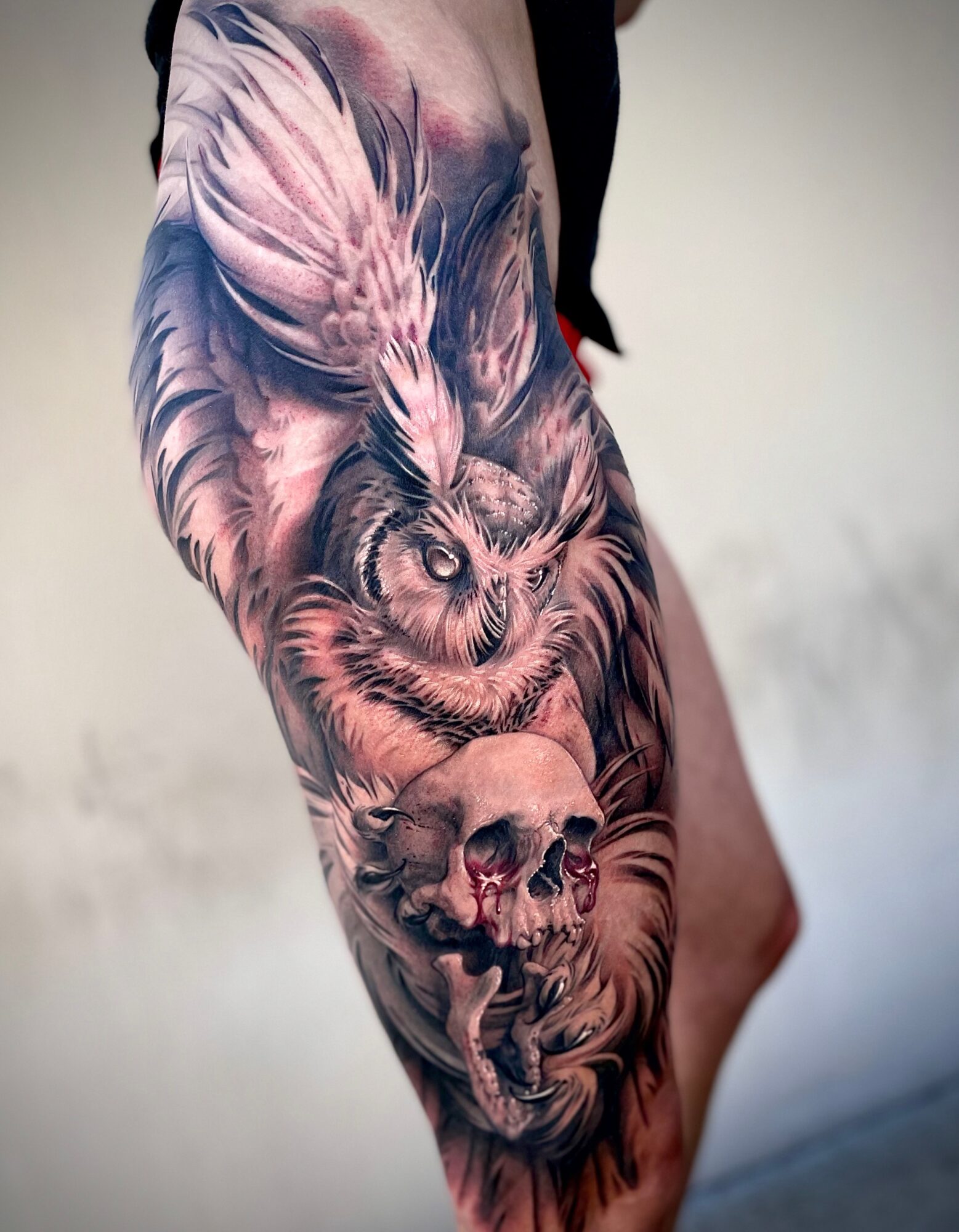 Tattoo of an owl with wings spread, perched above a skull, on a person's thigh, in black and gray shading.