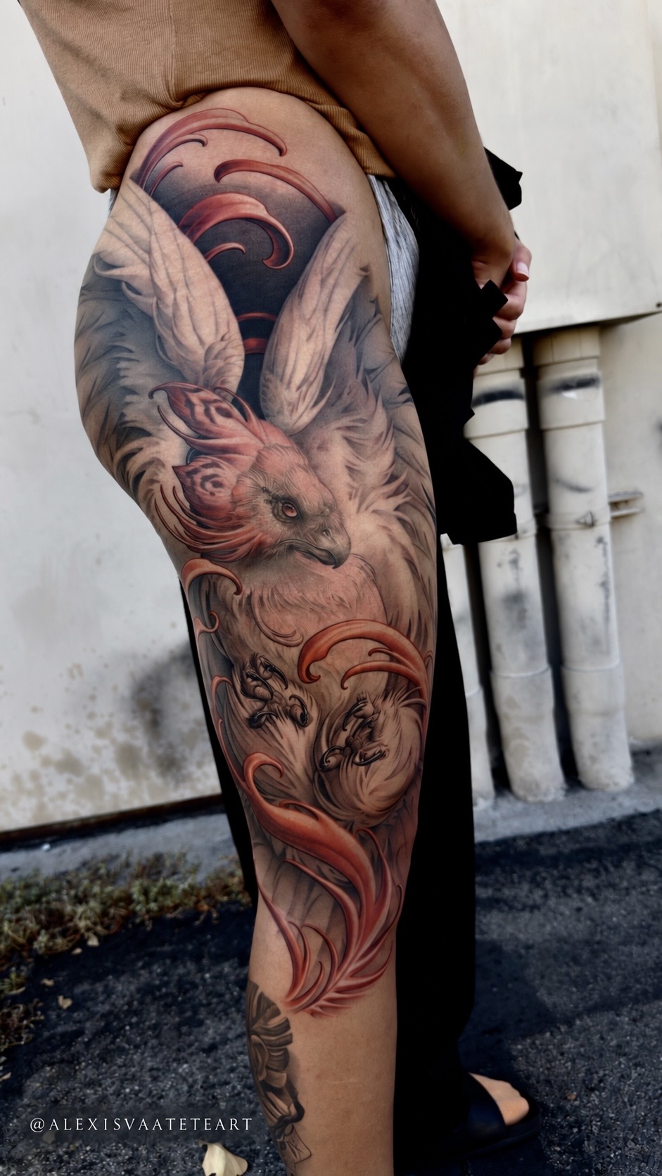 Tattooed thigh featuring a detailed angel with wings and flowing hair, with a background of clouds and sky.