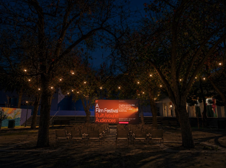 Night scene with string lights, trees, and a sign for a film festival, benches in foreground, dark sky.